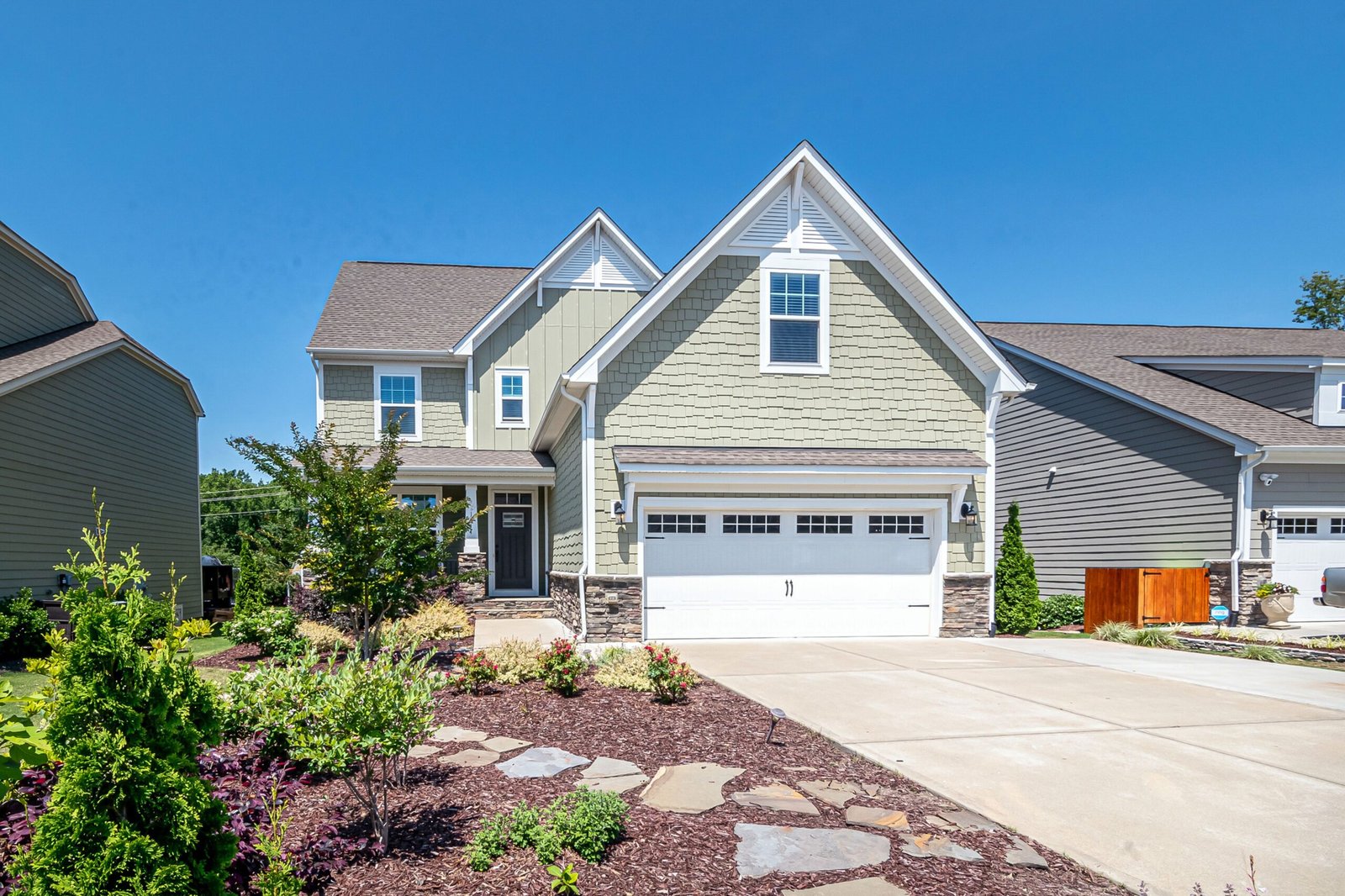 Charming suburban house with green facade, garage, and landscaped yard on a clear day.