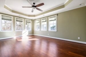 A bright and spacious empty room with large windows, hardwood floor, and ceiling fan.